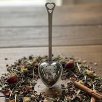 Heart-shaped tea spoon over a pile of dried herbs and a cinnamon stick on a wooden surface.
