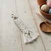 Macrame keychain with tassels on a wooden surface with yarn and knitting needles.