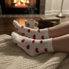 White socks with red hearts worn by a person sitting by a fireplace with a cup of coffee.