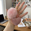 Hand holding a pink heart-shaped keychain with a fluffy pom-pom, desk and books in the background