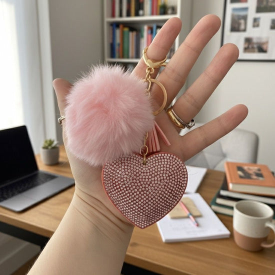 Hand holding a pink heart-shaped keychain with a fluffy pom-pom, desk and books in the background