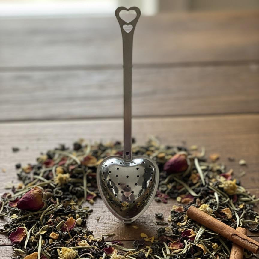 Heart-shaped tea spoon over a pile of dried herbs and a cinnamon stick on a wooden surface.