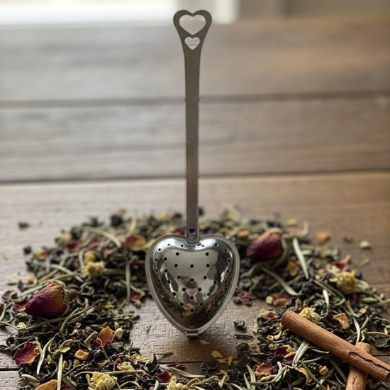 Heart-shaped tea spoon over a pile of dried herbs and a cinnamon stick on a wooden surface.