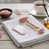 Pink facial roller and gua sha tool on a white towel with a cup of tea and rose petals in the background.