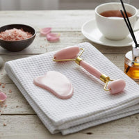 Pink facial roller and gua sha tool on a white towel with a cup of tea and rose petals in the background.