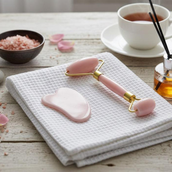 Pink facial roller and gua sha tool on a white towel with a cup of tea and rose petals in the background.